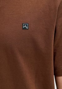 T-shirt en coton marron à manches courtes avec un petit logo noir en forme de carré et un motif de montagne sur la partie gauche de la poitrine.