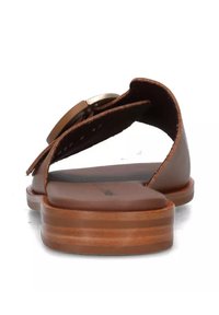 Sandal in pelle marrone con punta aperta, cinturini larghi e decorazione in metallo circolare. Suola piatta con una texture di legno marrone a strati.
