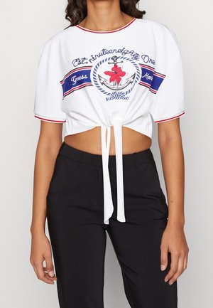 Femme portant un t-shirt court blanc avec un motif bleu marine, rouge et ancre noué à la taille, associé à un pantalon noir taille haute.