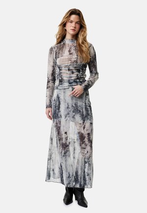 Kvinde iført en langærmet, højhalset, abstrakt grå og sort kjole med rynker, gennemsigtigt stof og sorte ankelstøvler, stående mod en hvid baggrund.