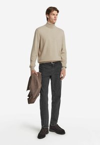 Mann trägt einen beigen Rollkragenpullover, dunkelgraue Jeans und dunkle Schuhe, hält eine braune Jacke in seiner linken Hand.