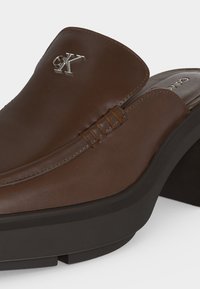 Καφέ δερμάτινο παπούτσι mule χωρίς χρώμα με μαύρη σόλα και ασημένιο λογότυπο Calvin Klein στο πάνω μπροστινό τμήμα.
