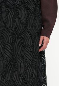 Μαύρο υφασμάτινο φόρεμα με υφή, που διαθέτει στρωματοποιημένα, κυματιστά σχέδια με βολάν, συμπληρωμένο από ένα μαρουνί μακρυμάνικο τοπ και ένα κοντινό πλάνο ενός χεριού.