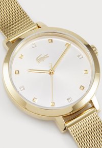 Reloj con correa de malla dorada y esfera plateada, con marcadores de hora cuadrados, un logo de Lacoste y dos manecillas doradas. Presenta piedras brillantes como acento.