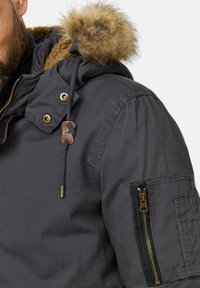 Chaqueta negra con capucha y cuello de piel, con detalles en dorado, bolsillos con cremallera y costuras decorativas. Fabricada con un material duradero.