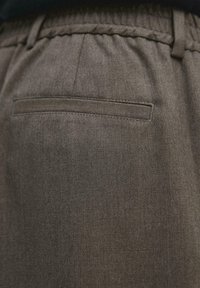 Gros plan d'un pantalon en tissu marron foncé montrant une poche passepoilée arrière et une ceinture élastique.