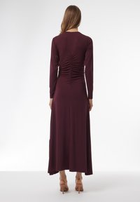 Largo vestido burdeos con mangas largas, detalle fruncido en la parte posterior ceñido al cuerpo y una falda fluida. El material parece suave y liso.