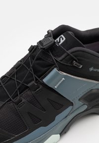 Chaussure de sport noire avec une tige en mesh texturé, des accents gris et bleu sarcelle, un système de laçage sécurisé et une semelle profilée amortissante.