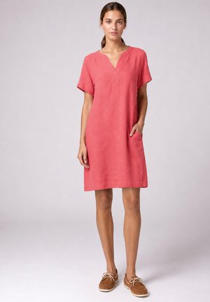 Femme portant une robe rose corail à manches courtes avec un col en V et une main dans la poche, associée à des chaussures bateau marron à lacets.