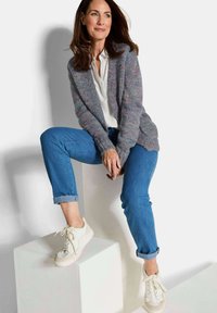 Vrouw met bruin haar zit op witte blokken, gekleed in een grijze gebreide cardigan, een wit shirt, blauwe opgerolde spijkerbroeken en witte sneakers.
