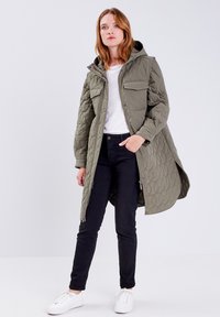 Quiltad olivgrön jacka med huva, fickor framtill och två bröstfickor; kombinerad med svarta byxor och vita sneakers som har en avslappnad passform.