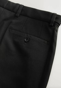 Čierne nohavice vyrobené z textúrovanej tkaniny, s plochým predným dielom, pútkami na opasok a zadnou vreckom so zapínaním na gombík.