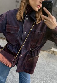 Vrouw die een spiegelselfie maakt terwijl ze een donkerdenim jack, blauwe jeans, parelringen draagt en een donkerpaarse schoudertas met een gouden schouderband draagt.