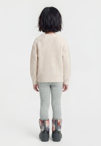 Παιδί με κοντά μαύρα μαλλιά στέκεται με την πλάτη, φορώντας ένα εκρού πλεκτό πουλόβερ, γκρι κολάν και πολύχρωμα χιονιά.