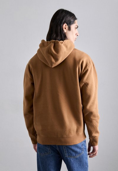 Sweatshirt à capuche marron en matériau doux, avec une coupe décontractée et un ourlet côtelé, associé à un jean en denim bleu.