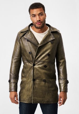 Olijfgroen leren trenchcoat met een dubbelzijdig ontwerp, grote revers, zwarte knopen en zijzakken, gedragen over een licht shirt.