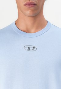 Nærbilde av en person som har på seg en lys blå t-skjorte med rund hals, med et lite sølvfarget ovalt emblem sentrert på brystet.