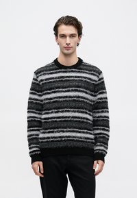 Young man wearing a black and gray fuzzy striped sweater and black pants, standing against a plain white background.