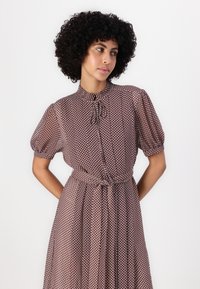 Rochie cu model maro, cu mâneci scurte tip puff, guler, detaliu cu decolteu în formă de cheie și o talie ajustată cu o curea.