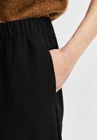 Pantalon noir avec une texture douce et une taille élastique, présentant une poche latérale et un gros plan d'une main reposant dans la poche.