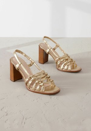Guldstrappy högklackade sandaler med en kraftig träklack, med ett vävt mönster på ovansidan och en slät, blank yta.