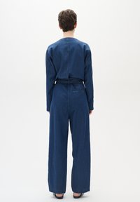 Denim overal v tmavě modré barvě s dlouhými rukávy, širokými nohavicemi a zavázaným pasem. Obsahuje zadní kapsu a hladkou texturu.