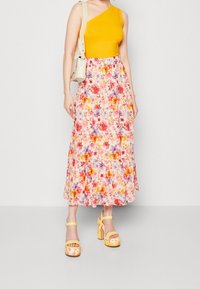 Frau trägt ein gelbes ärmelloses Oberteil, einen geblümten Midirock, passende geblümte Plateausandalen und eine beige Schultertasche.