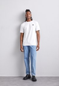 Vit kortärmad t-shirt med en svart logotyp, parat med ljusblå raka jeans och svarta snörskor mot en strukturerad grå bakgrund.