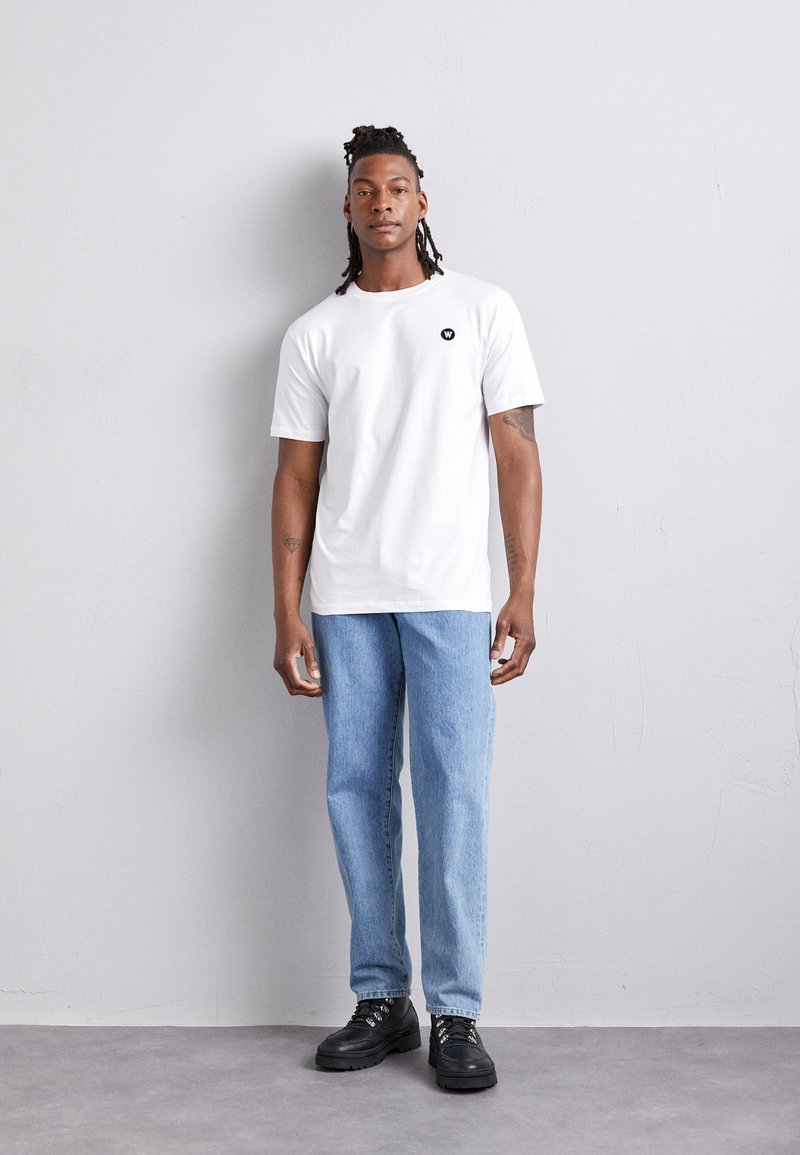 Vit kortärmad t-shirt med en svart logotyp, parat med ljusblå raka jeans och svarta snörskor mot en strukturerad grå bakgrund.