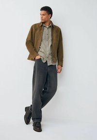 Brown suede jacket over a grey button-up shirt, paired with dark grey jeans and black lace-up shoes. Simple design with a relaxed fit.