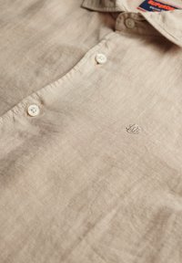 Beige linnen shirt met een kraaghalslijn, voorzien van twee knopen aan de voorkant en subtiele geborduurde details op de stof. Zachte textuur.