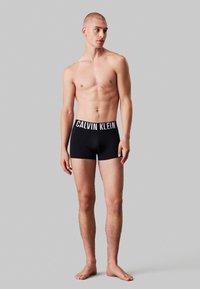 Schwarze Boxer-Shorts mit einem weißen, elastischen Bund mit Logo. Der Stoff wirkt glatt und eng anliegend. Das Modell steht barfuß vor einem neutralen Hintergrund.