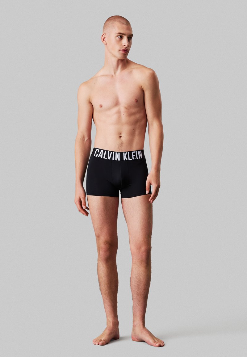 Schwarze Boxer-Shorts mit einem weißen, elastischen Bund mit Logo. Der Stoff wirkt glatt und eng anliegend. Das Modell steht barfuß vor einem neutralen Hintergrund.