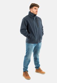 Veste légère bleu marine avec col haut et poche zippée, associée à un jean bleu clair et des bottes beiges. Texture lisse, coupe décontractée.