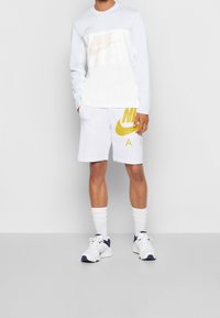 Bílá košile s dlouhým rukávem a síťovinou uprostřed, žluté logo Nike. Bílé šortky s velkým žlutým logem Nike. Bílé ponožky a tenisky.