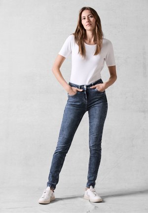 Jeans en denim bleu foncé avec une coupe slim, dotés de cinq poches et d'une fermeture éclair, assortis à un t-shirt blanc uni et des baskets blanches.