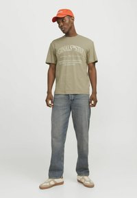 Olijfgroene katoenen t-shirt met witte grafische tekst, gecombineerd met lichtblauwe spijkerbroek. Oranje pet en beige sneakers met accenten.