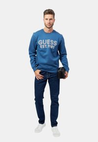 Felpa blu con il testo bianco "GUESS EST. 1981", abbinata a jeans blu scuro e sneakers bianche, mentre tiene in mano una piccola borsa nera.