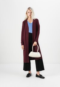 Lange bordeauxrode cardigan, losse pasvorm over een blauwe V-hals top, gecombineerd met zwarte wijde broek en een witte handtas. Zwarte schoenen maken de look af.