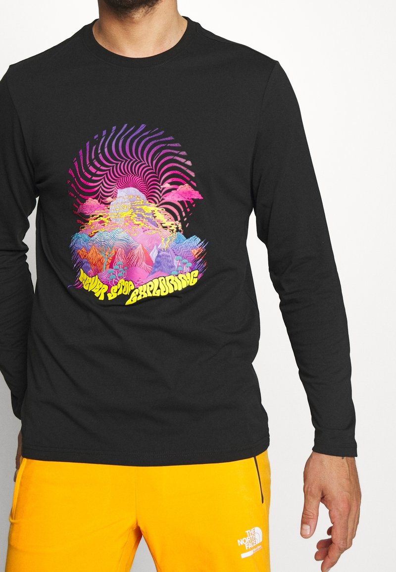 Muž v černém tričku s dlouhým rukávem, na kterém je barevný psychedelický design hor a nápis "Nikdy nepřestávej prozkoumávat", v kombinaci s jasně oranžovými kalhotami.
