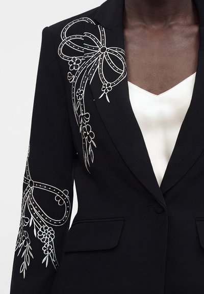 Μαύρο σακάκι με λευκή κεντητή διακόσμηση λουλουδιών και φιόγκων στον ώμο και το μανίκι, φορεμένο πάνω από λευκό σατέν τοπ με V-λαιμόκοψη.