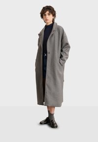 Abrigo largo gris de lana con cuello de solapa, cierre de un solo botón y bolsillos laterales. Lleva puesto sobre una parte superior oscura y pantalones cortos de mezclilla, combinado con zapatos tipo mocasín.