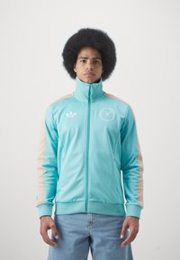 Blyantsjakke med lynlås i lys teal med orange striber langs ærmerne. Har høj krave og broderet logo på brystet. Blød stoftekstur.