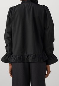 Vista posteriore di una persona che indossa una blouse nera a maniche lunghe con orlo e polsini arricciati, abbinata a pantaloni grigio scuro.