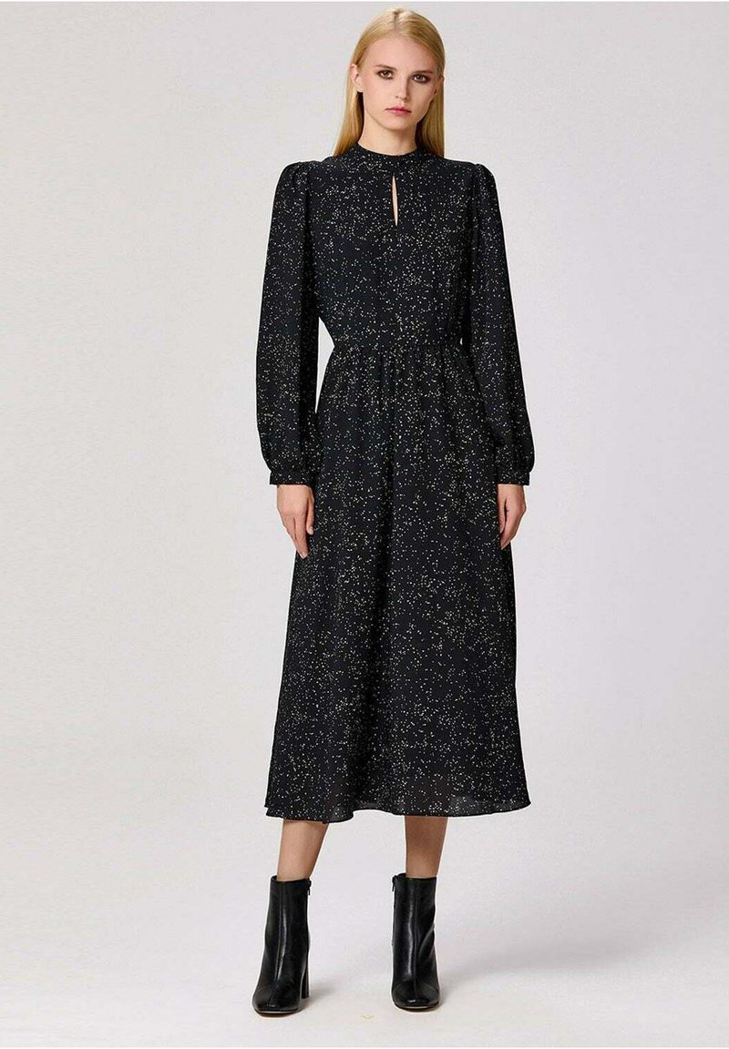 Robe noire à manches longues avec un décolleté en trou de serrure, taille ajustée et jupe fluide parsemée de paillettes dorées. Portée avec des bottines noires.
