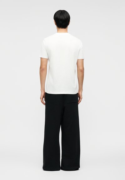 Άτομο με κοντά μαύρα μαλλιά, που φοράει απλό λευκό κοντομάνικο πουκάμισο και φαρδιά μαύρη παντελόνα, στέκεται με την πλάτη γυρισμένη σε έναν κενό φόντο.