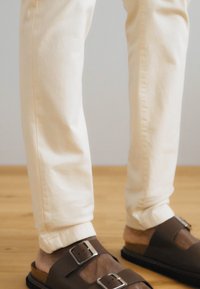 Un pantalon beige clair avec une coupe slim, associé à des sandales marron dotées de grandes boucles en argent et de semelles texturées. Texture de tissu lisse.