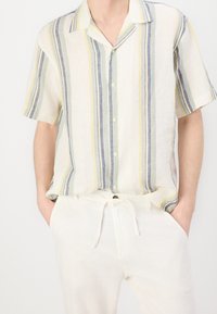 Kortärmad skjorta med knappar i ljust linne med vertikala ränder i blekgult, grönt och marinblått. Bärs med byxor i krämfärg.
