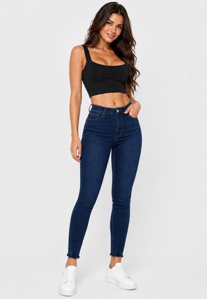 Frau mit langen dunklen Haaren, die ein schwarzes, bauchfreies Tanktop, hoch taillierte dunkelblaue Skinny-Jeans und weiße Sneaker trägt, steht mit einer Hand in der Tasche.