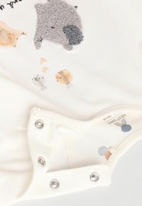 Bodysuit bébé blanc avec des appliqués d'animaux en gris, beige et couleurs douces. Possède des boutons-pressions en bas et une texture douce.